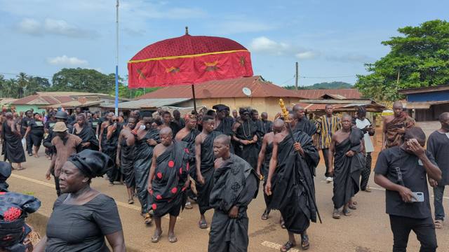 A procession of elders in black moves along the road beneath a red state umbrella.